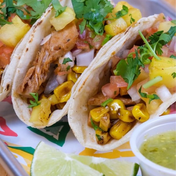 Three vibrant Taco Love tacos filled with meat, pineapple, corn, and cilantro, with lime wedges and salsa.