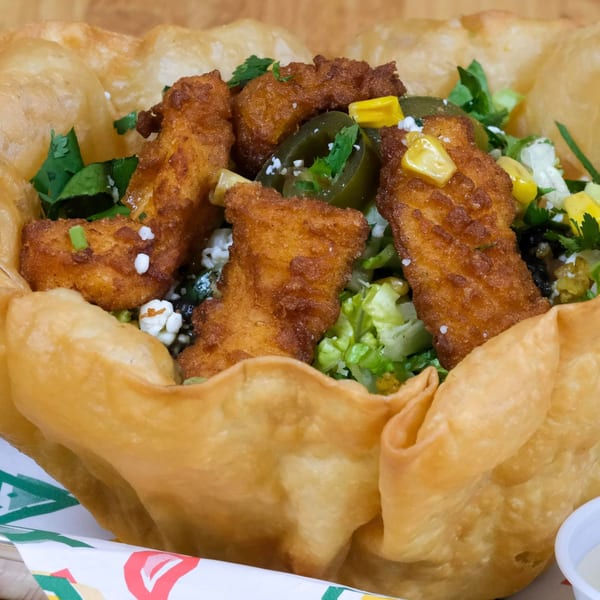 Crispy fried chicken pieces, corn, jalapeños, and lettuce in a large fried taco shell bowl.