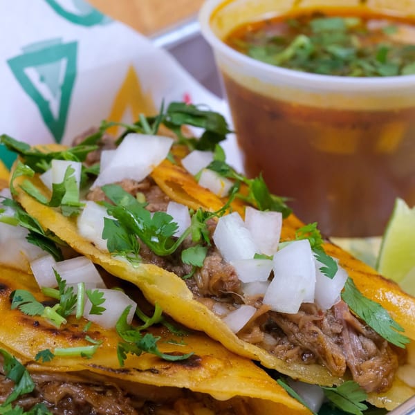 Three birria tacos with cilantro and diced onions, served with a side of consommé.