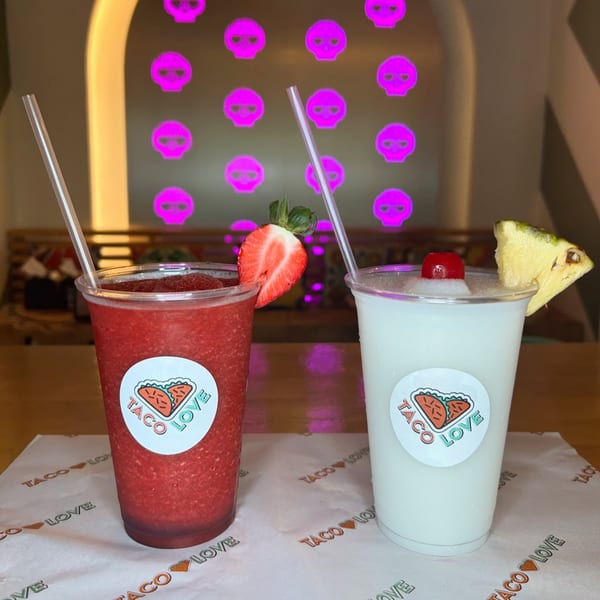 Two frozen drinks, one strawberry and one pina colada, with Taco Love logos and neon skull background.