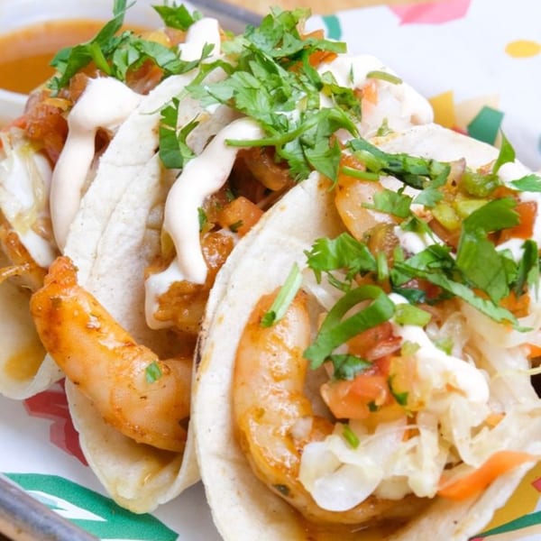 Three shrimp tacos with white sauce, salsa, and cilantro on a colorful paper liner.