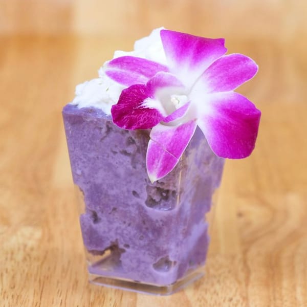 Purple dessert with white topping and a pink orchid in a clear glass on a wooden surface.